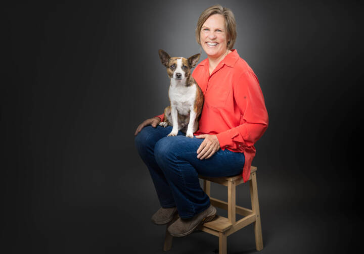 Rena Wells sitting in a chair with her dog on her lap against a grey background.