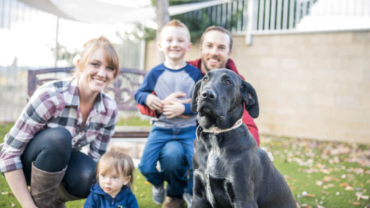 A family of a man, woman, and two children sitting outside with a black adoptable dog.
