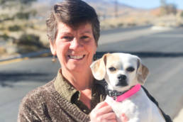 Photo of Kelley Bollen holding a small white dog, both are smiling at the camera, with beautiful snowcapped mountains behind them.
