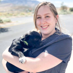 Photo of Olivia holding a black puppy and smiling, desert landscape behind her.