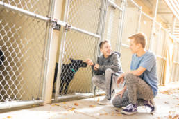 Two young men looking at a dog inside a kennel, smiling.